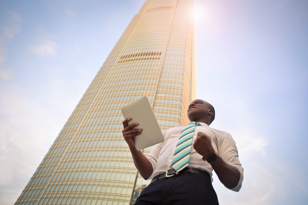 Man standing in front of tall building holding tablet who is really happy about seeing something on his screen